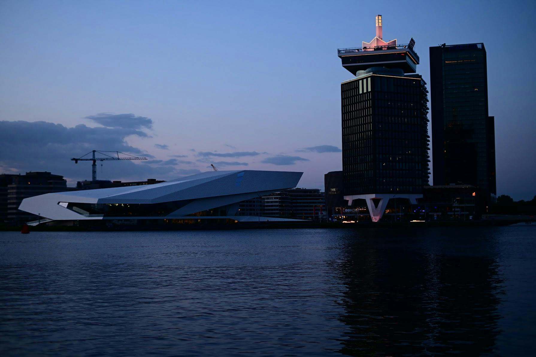 Picture of 3 buildings in focus during the blue hour, one large white building shaped as trapezoid and a large building that has a tower on it with three x, the amsterdam logo